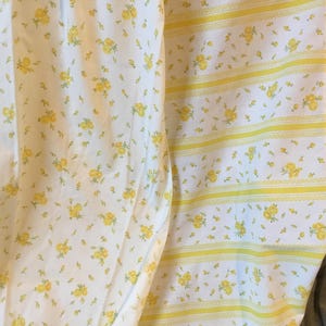 May include: A white fabric featuring a repeating pattern of small yellow flowers. One section displays a dense floral print, while the adjacent section features alternating vertical stripes of yellow floral patterns and thin yellow decorative bands.