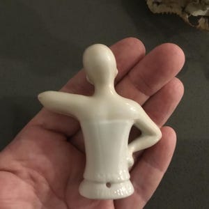 May include: A small, off-white porcelain doll torso with arms outstretched. The doll's back is visible, showing the smooth, curved form. The torso is held in a hand, with a dark background. The doll's form is simple and elegant.