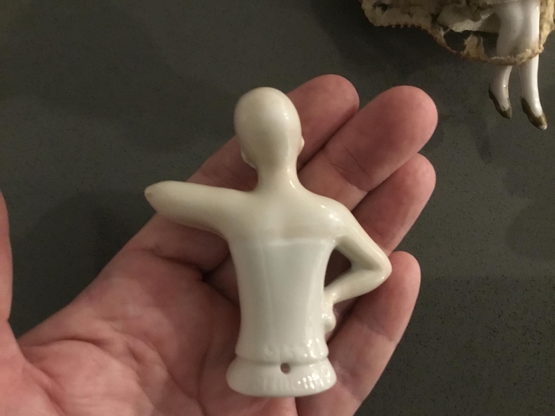 May include: A small, off-white porcelain doll torso with arms outstretched. The doll's back is visible, showing the smooth, curved form. The torso is held in a hand, with a dark background. The doll's form is simple and elegant.