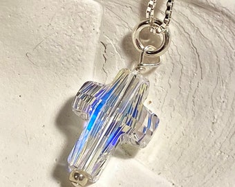 May include: A silver chain necklace with a crystal cross pendant. The cross is faceted and iridescent, reflecting light in shades of blue and white.