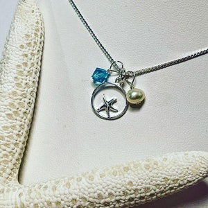 May include: A silver necklace with a starfish charm, a blue crystal charm, and a pearl charm. The necklace is on a white background.
