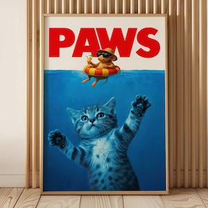 May include: A framed art print featuring a blue kitten reaching upwards towards a mouse in a life preserver. The word "PAWS" is printed in red at the top. The artwork is a parody of the movie "Jaws".