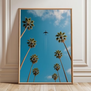 Op de afbeelding: Een ingelijste print met een helderblauwe lucht met wolken en hoge palmbomen. Een straalvliegtuig vliegt door de lucht en laat een witte condensstreep achter. Het kunstwerk wordt gepresenteerd in een houten lijst, leunend tegen een witte muur.