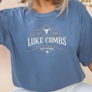 May include: A blue t-shirt with a vintage-style graphic design featuring the text "Luke Combs" and "The Legend" with a bull skull and the year 1990.