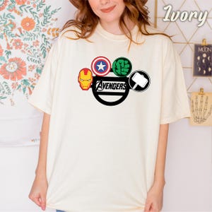 May include: Ivory-coloured t-shirt with the Avengers logo. The logo features Iron Man's mask, Captain America's shield, Hulk's fist, and Thor's hammer. The word "Avengers" is printed in a black, stylised font.