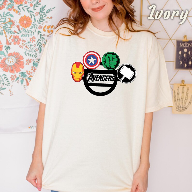 May include: Ivory-coloured t-shirt with the Avengers logo. The logo features Iron Man's mask, Captain America's shield, Hulk's fist, and Thor's hammer. The word "Avengers" is printed in a black, stylised font.