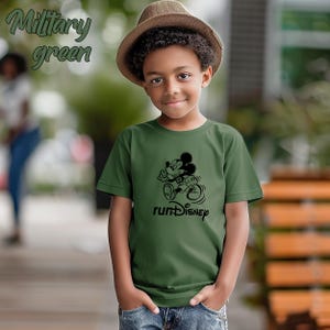 May include: A military green t-shirt featuring a black graphic of Mickey Mouse running with the text "runDisney". The shirt is worn by a young person wearing a brown hat and blue jeans.
