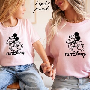 May include: Light pink t-shirts with a black graphic of Mickey Mouse running, with the text "runDisney" below. The shirts are worn by two people, one adult and one child. The words "light pink" are written in cursive in the top left corner.