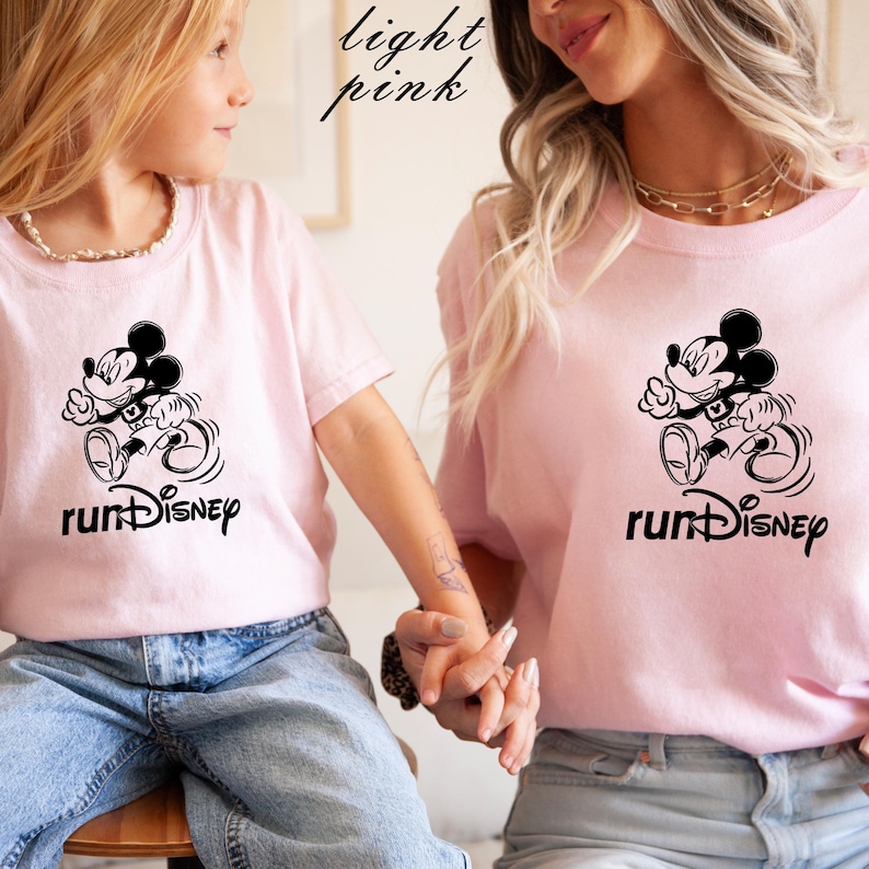 May include: Light pink t-shirts with a black graphic of Mickey Mouse running, with the text "runDisney" below. The shirts are worn by two people, one adult and one child. The words "light pink" are written in cursive in the top left corner.