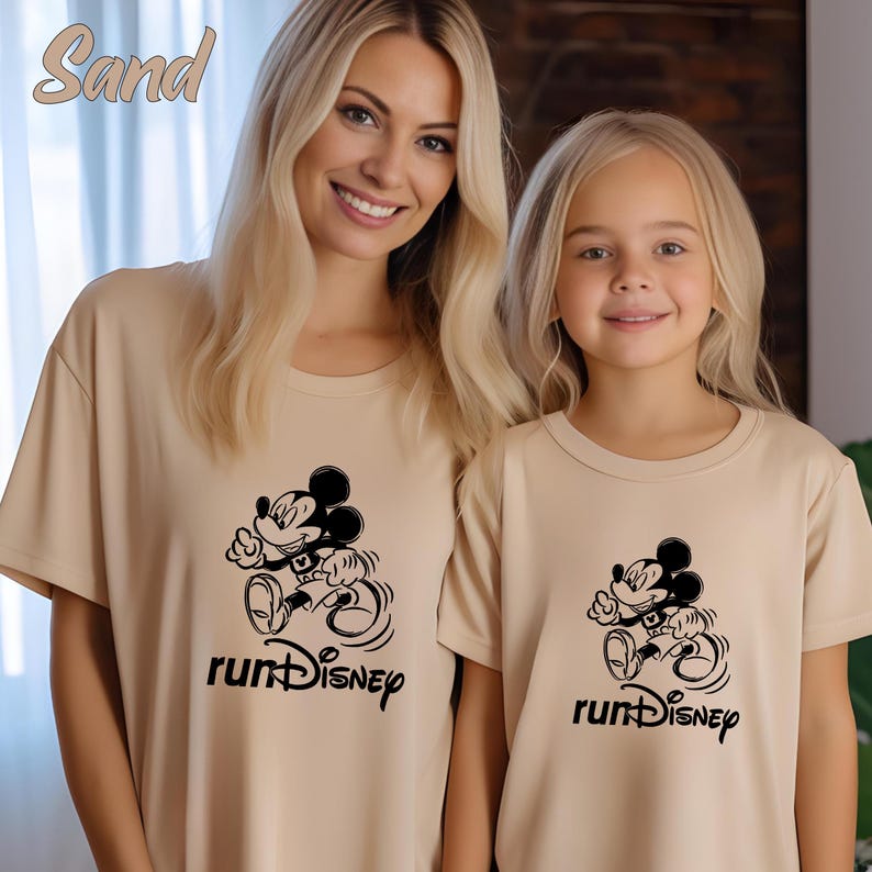 May include: Two beige t-shirts with a black Mickey Mouse graphic and the text "runDisney". The shirts are worn by two people. The word "Sand" is in the top left corner.