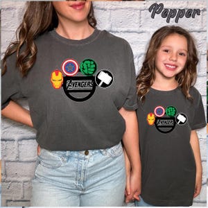 May include: Two charcoal grey t-shirts with the Avengers logo. The logo features Iron Man's mask, Captain America's shield, Hulk's fist, and Thor's hammer. The word "Avengers" is printed below the symbols in white.