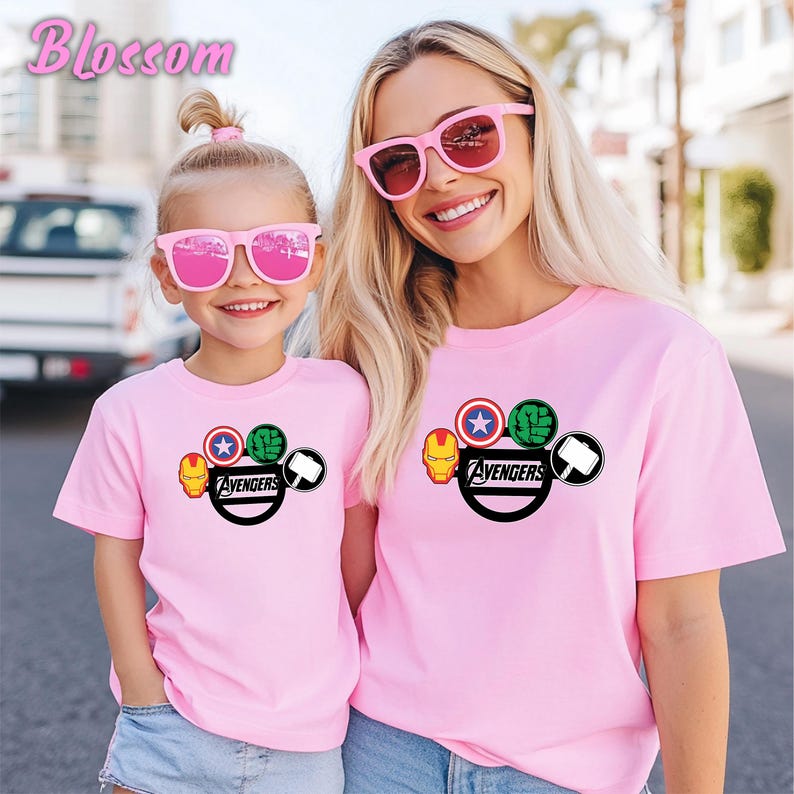 May include: Two pink t-shirts with the Avengers logo, featuring Iron Man, Captain America, Hulk, and Thor's hammer. The shirts are worn by a mother and child, both wearing pink sunglasses. The word "Blossom" is in the top left corner.