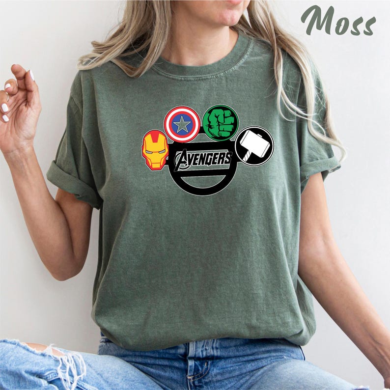 May include: Moss green t-shirt with the Avengers logo. The logo features Iron Man's mask, Captain America's shield, Hulk's fist, and Thor's hammer. The word "Avengers" is printed in white.