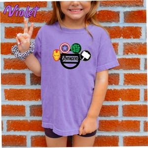 May include: A lilac t-shirt with the Avengers logo, including Iron Man, Captain America, Hulk, and Thor's hammer. The word "Avengers" is printed in black. The shirt is worn by a person in front of a brick wall.
