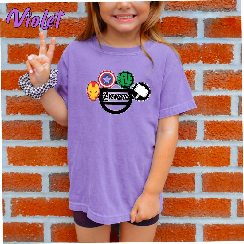 May include: A lilac t-shirt with the Avengers logo, including Iron Man, Captain America, Hulk, and Thor's hammer. The word "Avengers" is printed in black. The shirt is worn by a person in front of a brick wall.