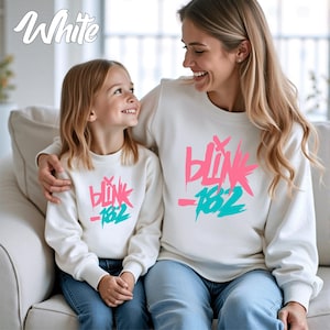 May include: White sweatshirts with the text "Blink 182" in pink and teal. A mother and child are wearing matching sweatshirts, sitting on a white couch. The word "White" is written in cursive in the upper left corner.