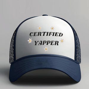 May include: A navy blue and white trucker hat with the words "CERTIFIED YAPPER" in black, surrounded by embroidered daisy accents. The hat has a mesh back and a curved brim.