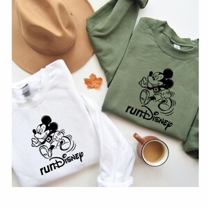 May include: Two white long-sleeved sweatshirts with a black Mickey Mouse graphic and the text "runDisney". One sweatshirt is on a green background and the other is on a brown background.