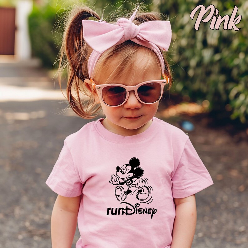 May include: A pink t-shirt featuring a black and white graphic of Mickey Mouse running, with the text "runDisney" below. The child is wearing pink sunglasses and a pink bow headband. The word "Pink" is in the top right corner.