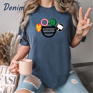 May include: Denim blue t-shirt with the Avengers logo, including Iron Man, Captain America, Hulk, and Thor's hammer. The word "Avengers" is in a black circle. A casual style t-shirt.