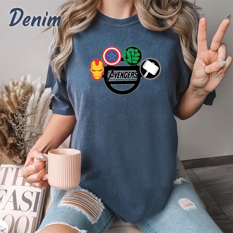 May include: Denim blue t-shirt with the Avengers logo, including Iron Man, Captain America, Hulk, and Thor's hammer. The word "Avengers" is in a black circle. A casual style t-shirt.