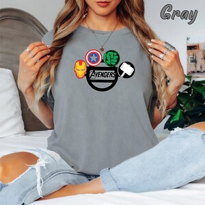 May include: A grey t-shirt displaying the Avengers logo, including Iron Man, Captain America, Hulk, and Thor's hammer. The word "Avengers" is printed in a black, stylised font. The shirt is a solid grey colour.