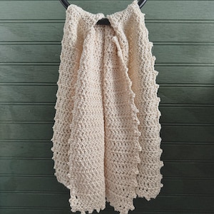 May include: A beige crocheted throw blanket with a scalloped edge hangs on a black metal ring.