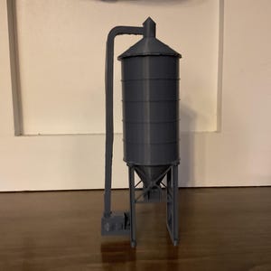 May include: A gray model of a grain silo, featuring a cylindrical storage tank with a conical roof. A vertical pipe extends from the top, curving to the side. The silo is supported by a metal frame, and sits on a wooden surface.