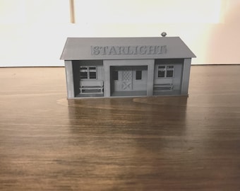 May include: A gray miniature building with the word "STARLIGHT" on the roof. The model has a small door, windows, and benches. The building is sitting on a wooden surface.