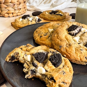 以下が含まれることがあります： 暗いお皿に盛られた、いくつかの大きなクッキーのクローズアップ。クッキーは、ダークチョコレートクッキーの破片、ホワイトチョコレートチップ、オレオのクッキーの破片が入ったゴールデンブラウンです。クッキーの1つは一口食べられています。