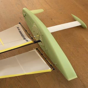 May include: A light green model airplane with a white wing and tail. The wings are made of a translucent material with yellow trim and black supports. The model is resting on a wooden surface.