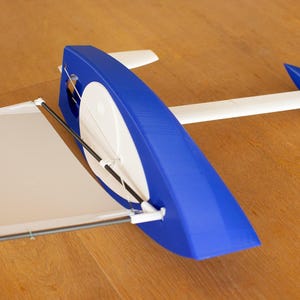 Può includere: Un modello di barca a vela blu e bianca con una vela bianca e uno scafo blu. Il modello è su una superficie di legno. La barca a vela ha un design aerodinamico, con un lungo albero bianco e una pinna blu.