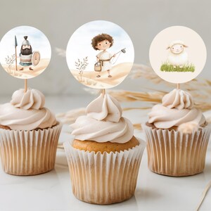 May include: Three cupcakes with white frosting topped with round paper circles featuring illustrations of a shepherd, a lamb, and a man with a spear. The cupcakes are on a white surface with dried grass.