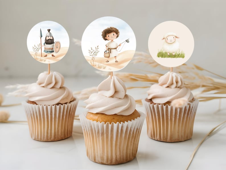 May include: Three cupcakes with white frosting topped with round paper circles featuring illustrations of a shepherd, a lamb, and a man with a spear. The cupcakes are on a white surface with dried grass.