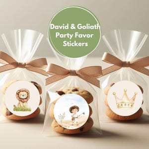 May include: Three clear cellophane bags with brown ribbon ties, each containing cookies and a round sticker. The stickers feature illustrations of a lion, a boy with a slingshot, and a crown. The green circle sticker on top reads "David & Goliath Party Favor Stickers".
