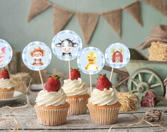Op de afbeelding: Cupcakes gegarneerd met aardbeien en witte glazuur, versierd met toppers met boerderijdieren en boeren. De toppers tonen een varken, boer, koe, eendje en schuur, tegen een blauwe gingham achtergrond.