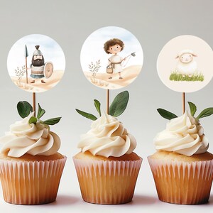 May include: Three cupcakes with white frosting topped with round paper cupcake toppers featuring illustrations of a shepherd, a lamb, and a warrior.