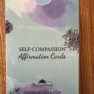 A box of self-compassion affirmation cards with a watercolor background in shades of purple and green. The box has the words 'Self-Compassion Affirmation Cards' printed in black text. The bottom of the box has the words 'True Embrace Therapy PLLC' and 'TANGELA R. MILLS, LCSW' printed in black text.