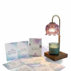 May include: A self-care set featuring affirmation cards, a candle, and a decorative lamp. The cards have inspirational quotes. The lamp has a pink glass shade and a gold-colored metal frame. The candle is in a clear glass holder.
