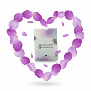 May include: A deck of "Self-Compassion Affirmation Cards" is centered within a heart shape made of purple flower petals. The card deck has a light green background with the text "Self-Compassion Affirmation Cards". The petals are various shades of purple.