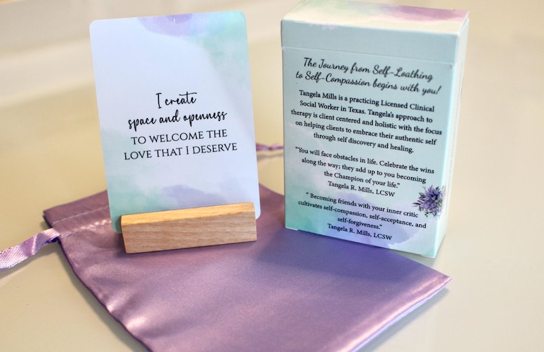 May include: A set of self-help cards with the text "I create space and openness to welcome the love that I deserve." The cards are displayed with a wooden stand, a satin pouch, and a box with text about self-compassion.