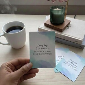 May include: A white mug filled with coffee sits next to a stack of books and a lit candle in a green glass holder. A hand holds a card with the text "Every day I am blooming INTO MY BEST SELF UNAPOLOGETICALLY."