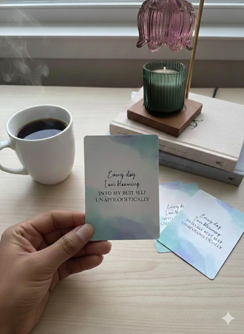 May include: A white mug filled with coffee sits next to a stack of books and a lit candle in a green glass holder. A hand holds a card with the text "Every day I am blooming INTO MY BEST SELF UNAPOLOGETICALLY."