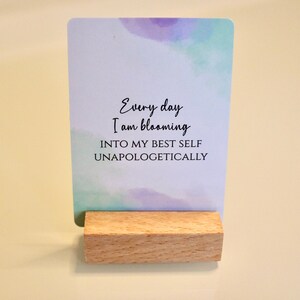May include: A rectangular card with watercolor-style blue and purple accents, held by a small wooden block. The card features the text "Every day I am blooming INTO MY BEST SELF UNAPOLOGETICALLY" in a handwritten font.