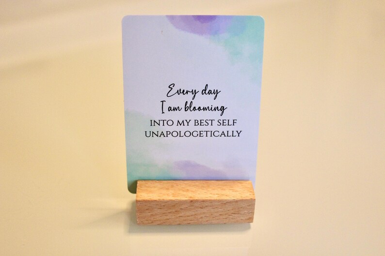 May include: A rectangular card with watercolor-style blue and purple accents, held by a small wooden block. The card features the text "Every day I am blooming INTO MY BEST SELF UNAPOLOGETICALLY" in a handwritten font.