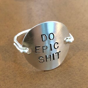 May include: A silver ring with a round, flat, silver disc that says "DO EPIC SHIT" in all caps.