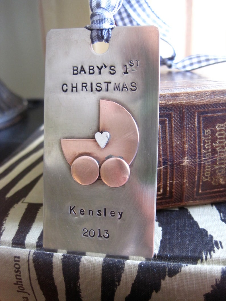 以下が含まれることがあります： 「BABY'S 1ST CHRISTMAS」と「Kensley 2013」の文字が刻まれた銀製のオーナメント。オーナメントには、白いハートがついた銅色のベビーカーが描かれている。