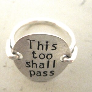 May include: A silver ring with a round charm that reads "This too shall pass".