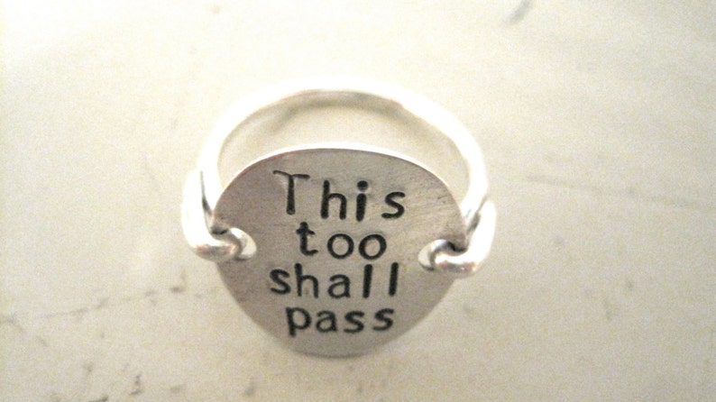 May include: A silver ring with a round charm that reads "This too shall pass".