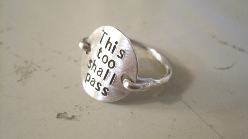 May include: Silver ring with a round charm that says "This too shall pass".